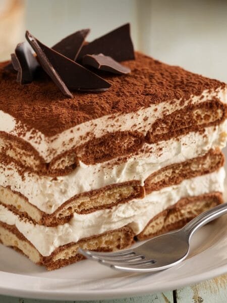 A slice of tiramisu with cocoa powder and chocolate shavings on a white plate.