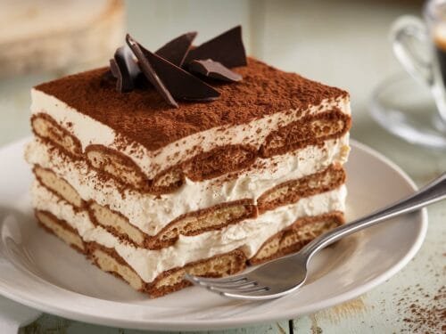 A slice of tiramisu with cocoa powder and chocolate shavings on a white plate.